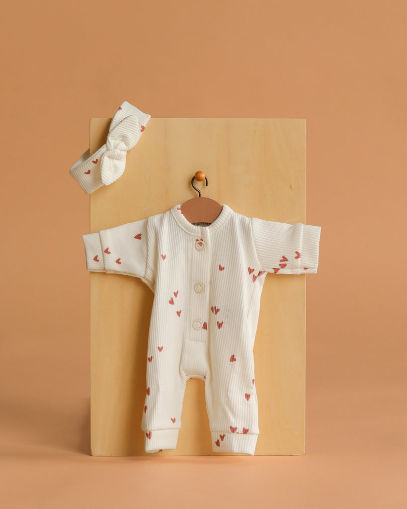 The Minikane 11" Doll Clothing set, featuring the Lili Jumpsuit & Headband in Ribbed Knit - Pink Hearts, is displayed on a wooden board against a light brown background, perfect as stylish doll clothes.