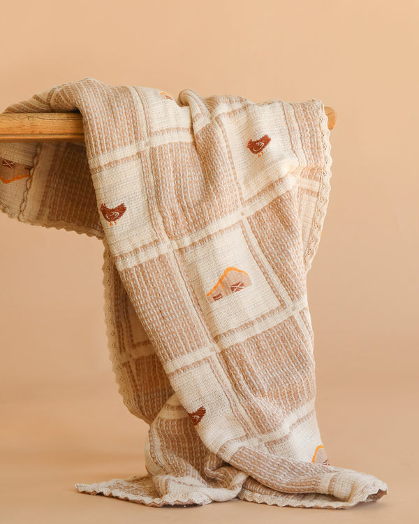 The Organic Cotton Baby Blanket - Farm features a cream and beige checkered pattern, crochet trim, and small brown embroidered chickens, draped over a wooden surface with a plain beige background.