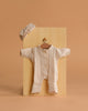 The Minikane 11″ Doll Clothing set features the Lili Jumpsuit & Headband in soft ecru cotton gauze, beautifully crafted in France. The jumpsuit with buttons is displayed on a small hanger, and the matching knotted headband rests above it—all set against a neutral tan background and perfectly sized for Minikane Dolls.