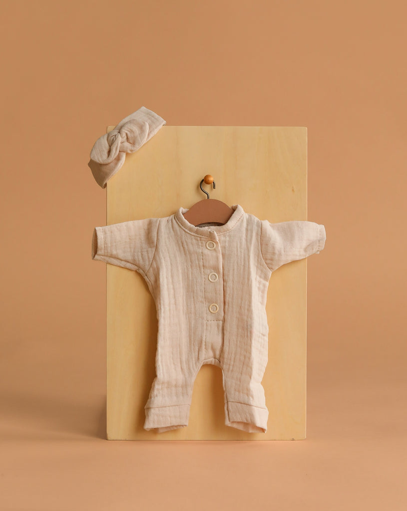 The Minikane 11″ Doll Clothing set features the Lili Jumpsuit & Headband in soft ecru cotton gauze, beautifully crafted in France. The jumpsuit with buttons is displayed on a small hanger, and the matching knotted headband rests above it—all set against a neutral tan background and perfectly sized for Minikane Dolls.