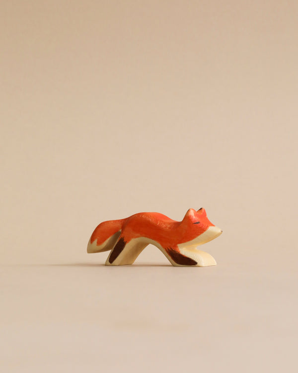 A small, hand-painted Holzwald Small Fox figurine in a leaping pose, placed against a plain, light beige background. The fox is predominantly orange with white details. This high-quality toy