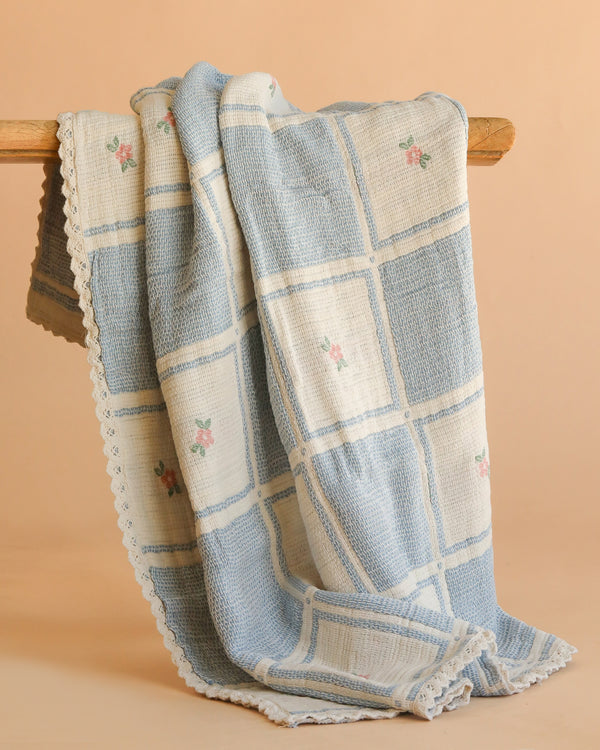 The Organic Cotton Baby Blanket - Spring Bloom features a blue and white checkered pattern, small pink embroidered flowers, and a scalloped edge, shown draped over a wooden rod against a beige background.