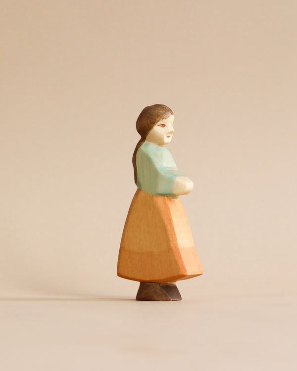A Handmade Holzwald Goose Girl figurine of a woman with a pastel blue shawl over a peach dress, colored with natural dyes, standing against a plain beige background.
