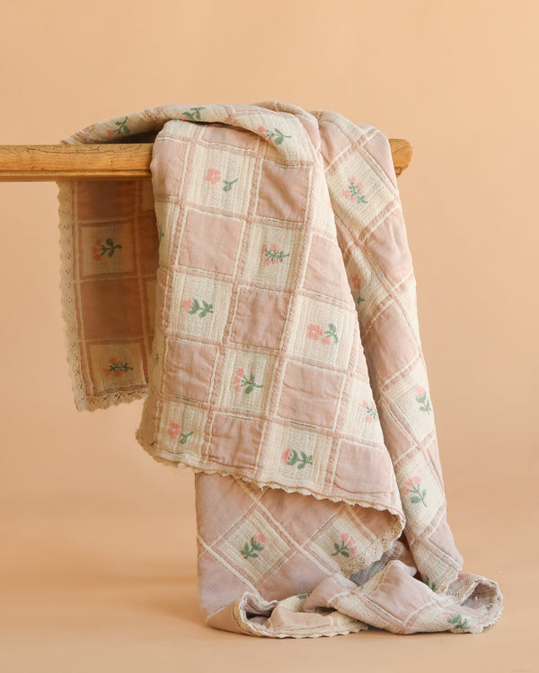 The Organic Cotton Baby Blanket - Fleur features a pastel pink and cream checkered pattern with small floral designs. Crafted in Portugal from organic cotton, this cozy blanket is displayed over wood with a beige background.