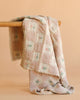 The Organic Cotton Baby Blanket - Fleur features a pastel pink and cream checkered pattern with small floral designs. Crafted in Portugal from organic cotton, this cozy blanket is displayed over wood with a beige background.