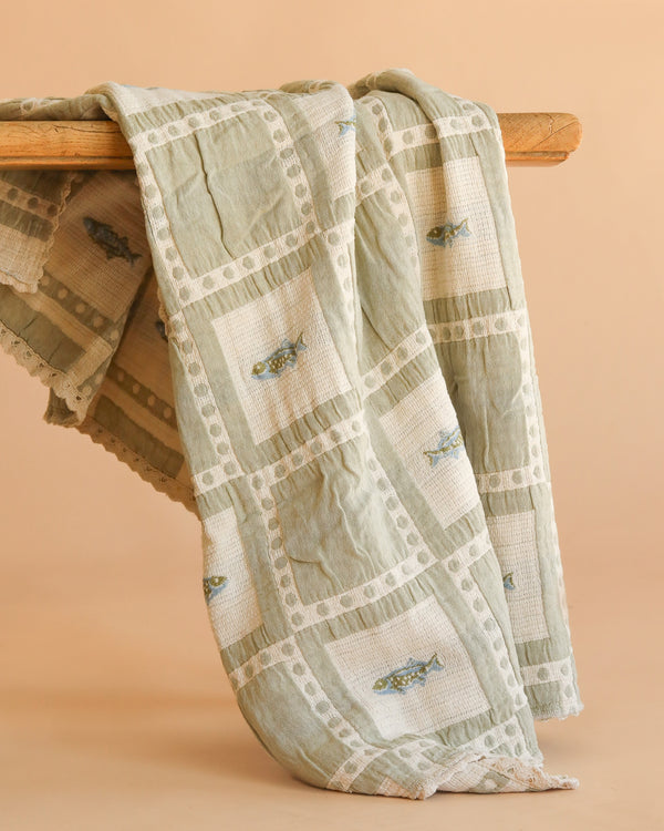 The Organic Cotton Baby Blanket - Fishing, in light green and cream with embroidered fish designs, is made in Portugal from organic cotton and hangs draped over a wooden rod against a beige background.