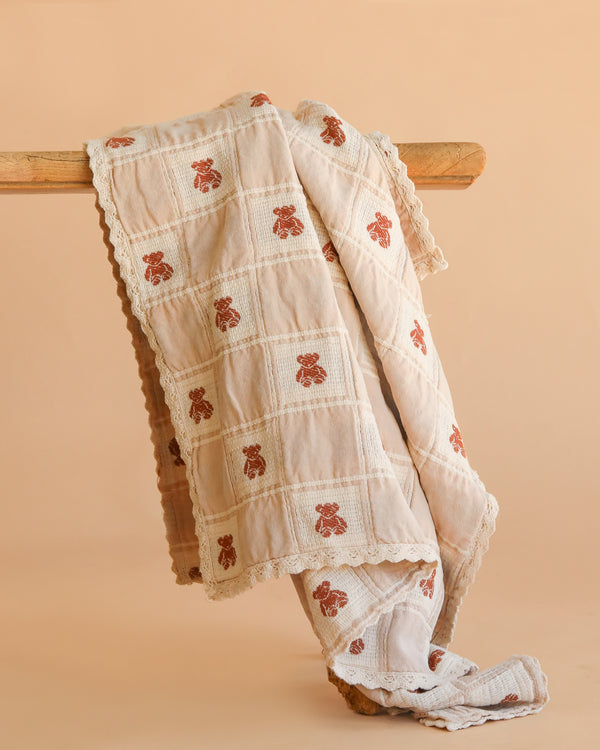 The Organic Cotton Baby Blanket - Teddy, featuring a cream color, checkered pattern, brown teddy bear motifs, and lace trim, is displayed draped over a wooden rod against a plain beige background.