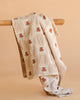 The Organic Cotton Baby Blanket - Teddy, featuring a cream color, checkered pattern, brown teddy bear motifs, and lace trim, is displayed draped over a wooden rod against a plain beige background.