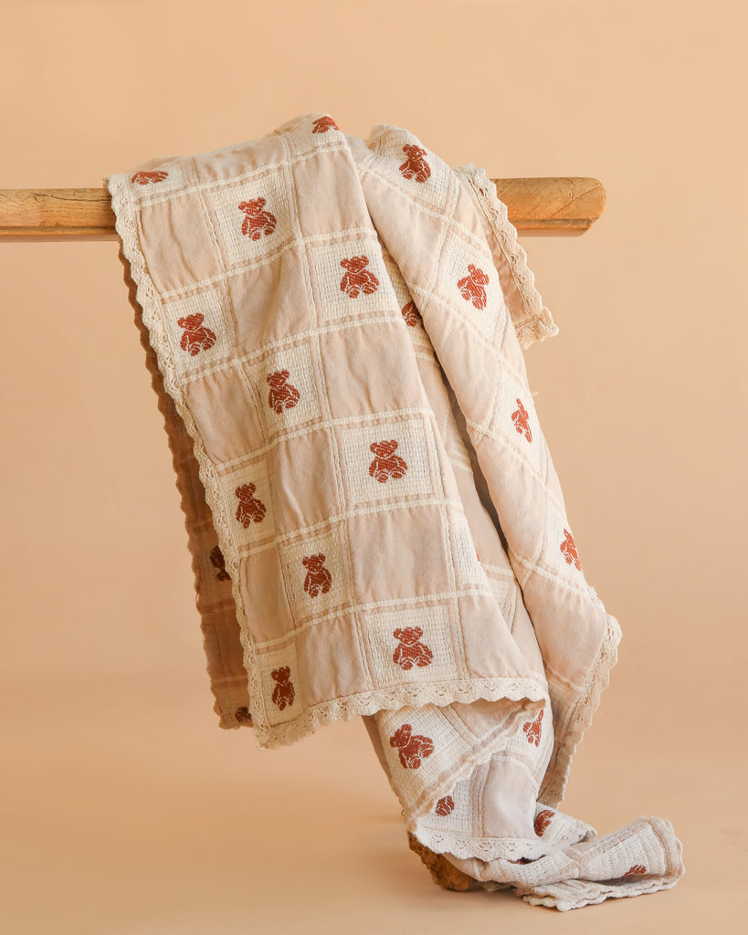 The Organic Cotton Baby Blanket - Teddy, featuring a cream color, checkered pattern, brown teddy bear motifs, and lace trim, is displayed draped over a wooden rod against a plain beige background.