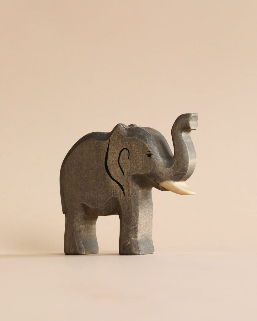 A Handmade Holzwald Elephant sculpture with raised trunk and ivory tusks, positioned against a plain, light beige background. This high-quality elephant has visible grain textures, giving it a rustic appearance.