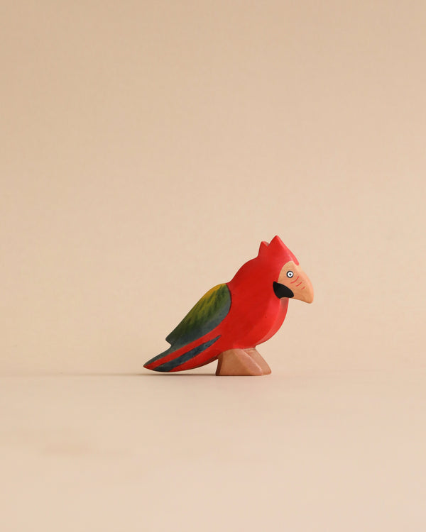 A colorful, high-quality Handmade Holzwald Parrot Bird figurine painted red with green and blue details, standing on a small brown base against a pale beige background.