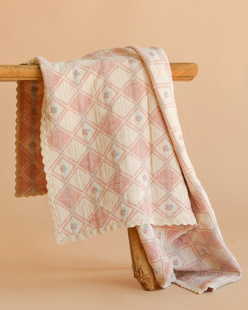 The Organic Cotton Baby Blanket - Bouquet, in soft pink and cream with a diamond pattern and scalloped edges, is draped over a rustic wooden beam against a plain light peach background.