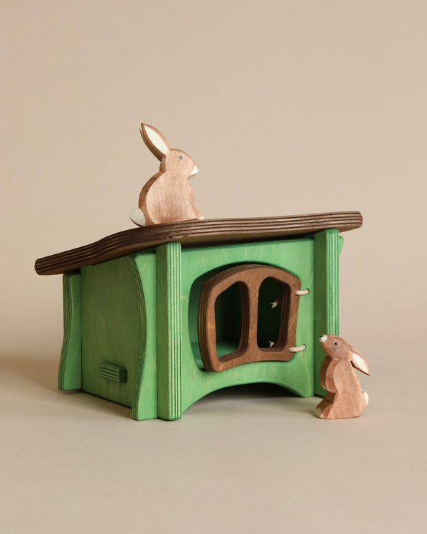 The Ostheimer Rabbit Hutch, painted green with a dark brown roof, designed for imaginative play, featuring cutout windows and a door. Two small wooden rabbits, one on the roof and another near the door.
