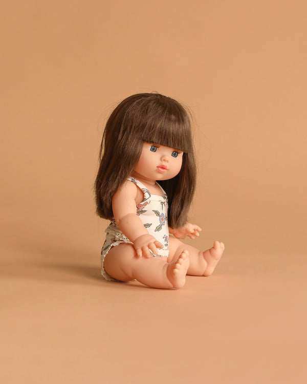 The Minikane Baby Doll (13") - Cherry with Clothing features straight brown hair and bangs, wearing a floral outfit. Seated on a matching light brown background and facing slightly right, this anatomically correct doll inspires imaginative play.