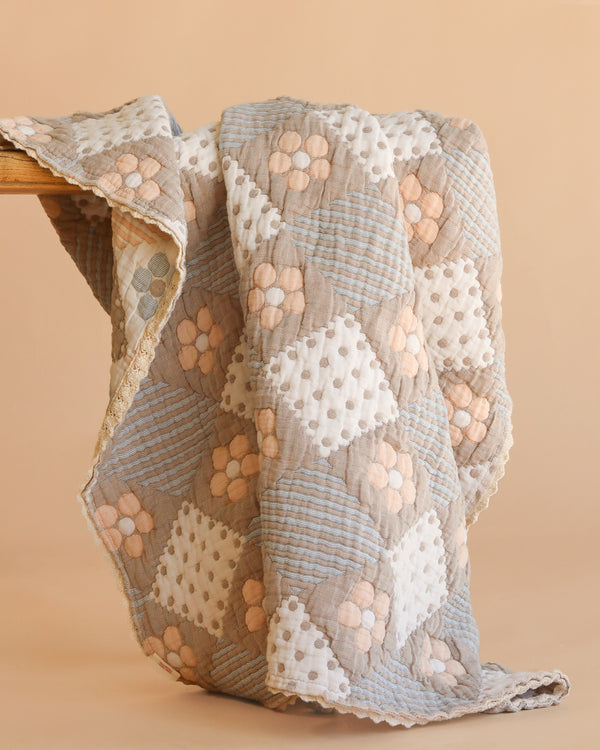 The Organic Cotton Baby Blanket - Prairie, handmade with a patchwork design, features soft pastel hues like beige, light blue, and white, accented by floral and polka dot patterns. The cozy quilt is draped over a wooden surface.