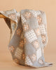 The Organic Cotton Baby Blanket - Prairie, handmade with a patchwork design, features soft pastel hues like beige, light blue, and white, accented by floral and polka dot patterns. The cozy quilt is draped over a wooden surface.