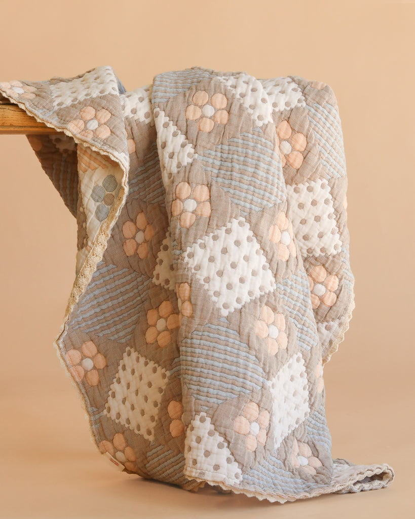 The Organic Cotton Baby Blanket - Prairie, handmade with a patchwork design, features soft pastel hues like beige, light blue, and white, accented by floral and polka dot patterns. The cozy quilt is draped over a wooden surface.