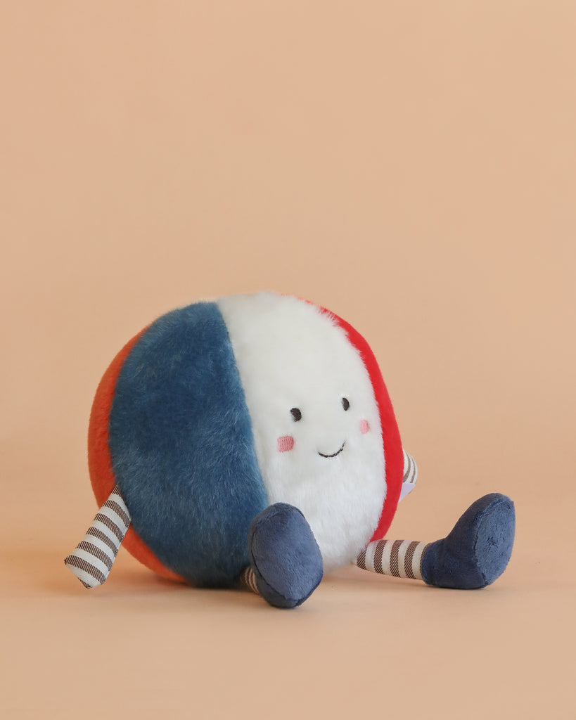 The Mon Ami Beach Ball Plush Toy, featuring blue, white, and red segments with a smiling face and striped arms and legs, sits on a peach-colored background.