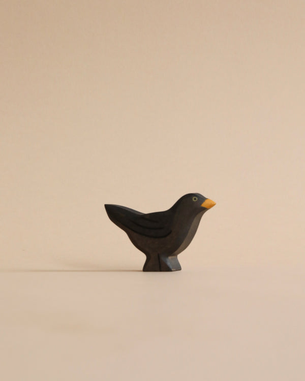 A small, simplistic Handmade Holzwald Blackbird sculpture, predominantly black with a yellow beak, crafted from high-quality wood, standing upright on a light tan surface against a matching background.