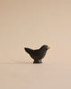 A small, simplistic Handmade Holzwald Blackbird sculpture, predominantly black with a yellow beak, crafted from high-quality wood, standing upright on a light tan surface against a matching background.