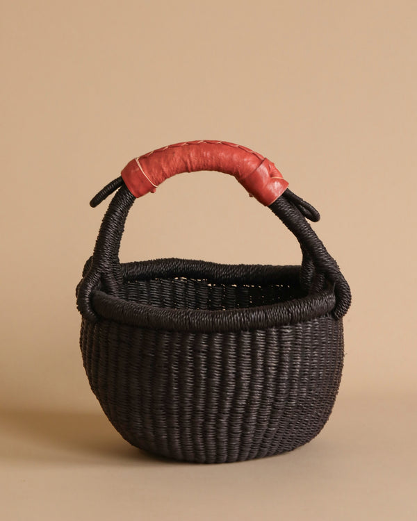 The Handmade Explore Basket is a round, dark woven basket made from elephant grass, featuring a sturdy handle wrapped in red material. Hand-made in Bolgatanga, Ghana, it boasts a tightly braided texture and is displayed against a plain beige background.