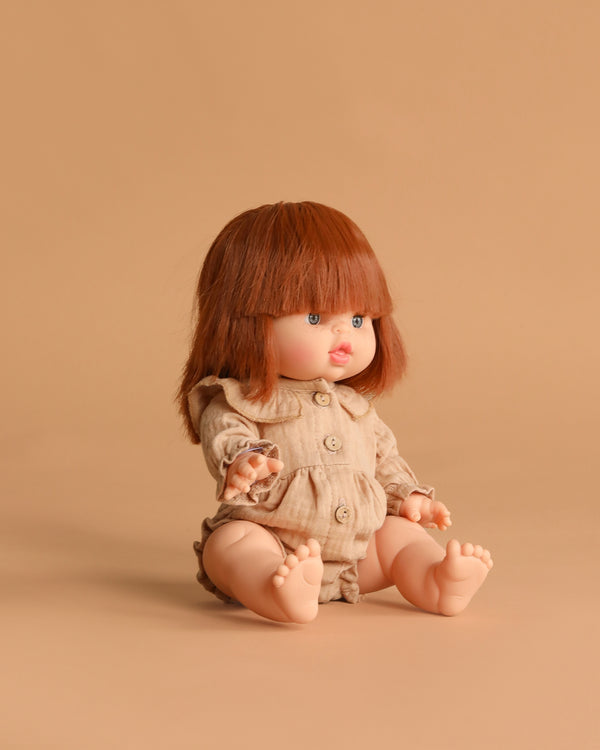 The Minikane Baby Doll (13") - Capucine with Clothing, featuring brown hair and bangs in a beige romper, sits barefoot on a matching beige background, facing slightly left.