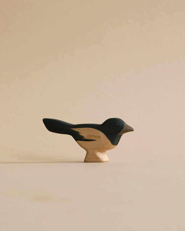 A Handmade Holzwald Magpie figurine, painted black and beige, stands against a pale beige background. The figure is not only a sustainable toy but also simplistic yet detailed, capturing the essence of a small bird.