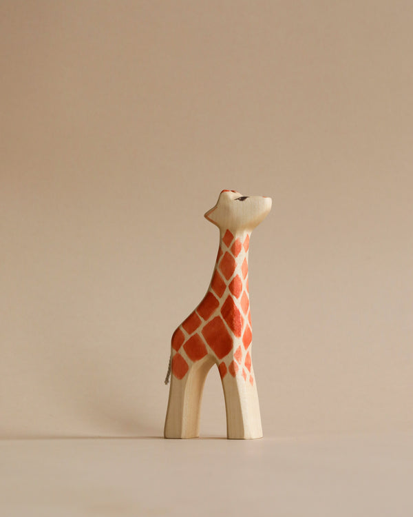 A sustainable Handmade Holzwald Giraffe - Small figurine painted with orange and white patterns stands against a neutral beige background. The giraffe is depicted in a simple, stylized form with a peaceful expression.