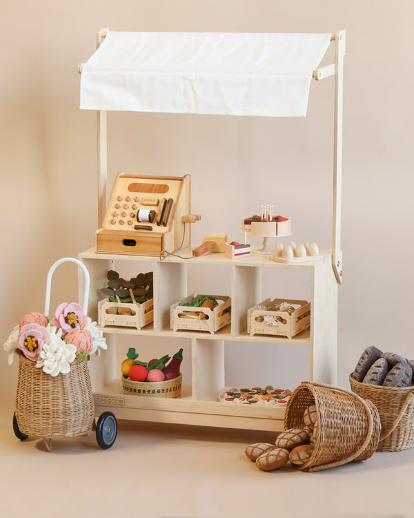 The Milton & Goose Market Stand is a stylish heirloom-quality wooden setup with a cash register, baskets of faux flowers and fruits, and various play food items on shelves, against a neutral background.