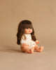 The Minikane Baby Doll (13") - Cherry with Clothing and Stroller Set sits on a neutral beige background, dressed in white with long brown hair and bangs, legs outstretched.