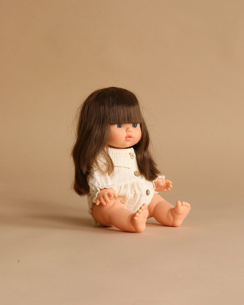 The Minikane Baby Doll (13") - Cherry with Clothing and Stroller Set sits on a neutral beige background, dressed in white with long brown hair and bangs, legs outstretched.
