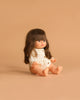 The Minikane Baby Doll (13") - Cherry with Clothing, featuring long brown hair and bangs in a cream button-up romper, sits on a light brown surface against a matching background.