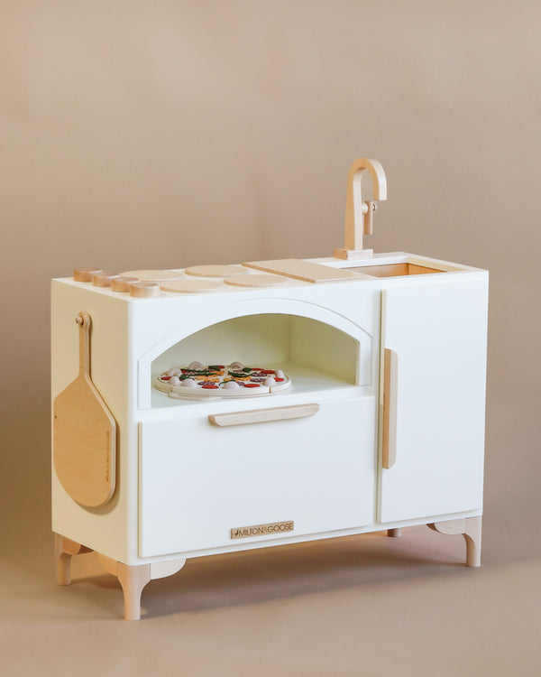 A Milton & Goose play kitchen set against a beige background, featuring a stove, an oven, and a sink, with interactive educational toys like a pizza-making set and a spatula on top. The kitchen is made in the USA.