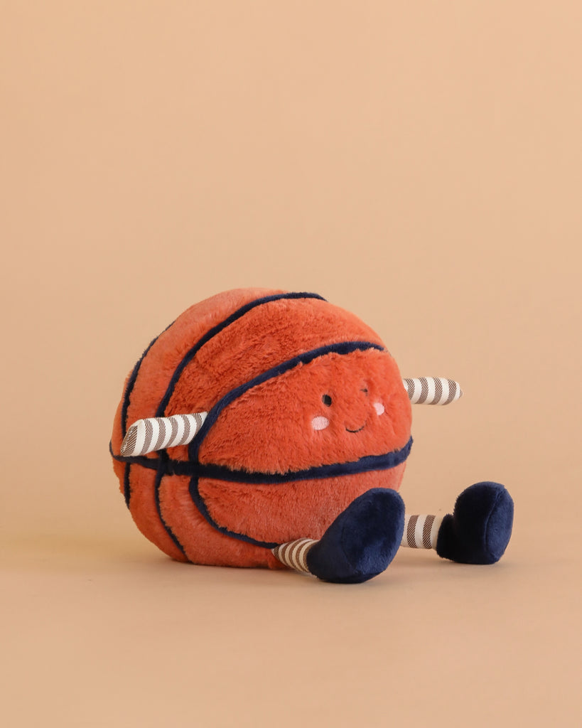 The Mon Ami Barclay Basketball Plush Toy, with a smiling face, striped arms and legs in blue shoes, sits on a beige surface—an adorable cuddle buddy and perfect baby gift.