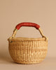The Easter Basket Gift Set features a round, woven straw basket with a sturdy handle wrapped in red material, displayed on a plain beige background.