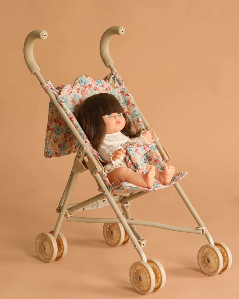 The Minikane Baby Doll (13") - Cherry with Clothing and Stroller Set features a vintage-style doll in a floral dress seated in a beige stroller with floral fabric accents, all against a plain beige background.