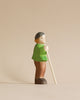 A handmade Holzwald Shepherd figurine of an elderly man with a walking stick, painted in green and brown, standing against a plain beige background from the Holzwald Brand.