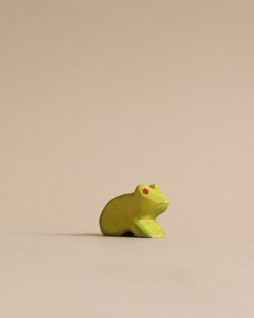 A small, yellow Handmade Holzwald Frog with red eyes, positioned centrally against a plain, light beige background treated with high-quality oil.