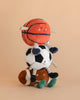 The Mon Ami Sports Ball Set features three plush toys—a football, basketball, and soccer ball—each with smiling faces and striped arms and legs, stacked atop one another against a plain beige background.