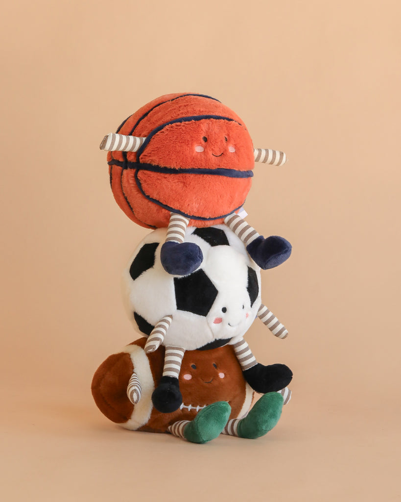 The Mon Ami Sports Ball Set features three plush toys—a football, basketball, and soccer ball—each with smiling faces and striped arms and legs, stacked atop one another against a plain beige background.