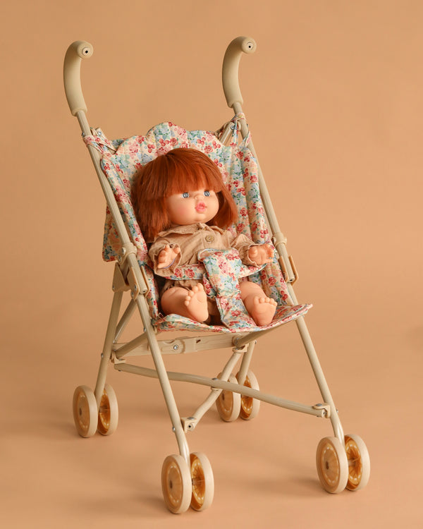 The Minikane Baby Doll (13") - Capucine with Clothing and Stroller Set features a red-haired doll in a beige outfit, seated in a floral-patterned stroller with beige wheels, set against a light brown background.