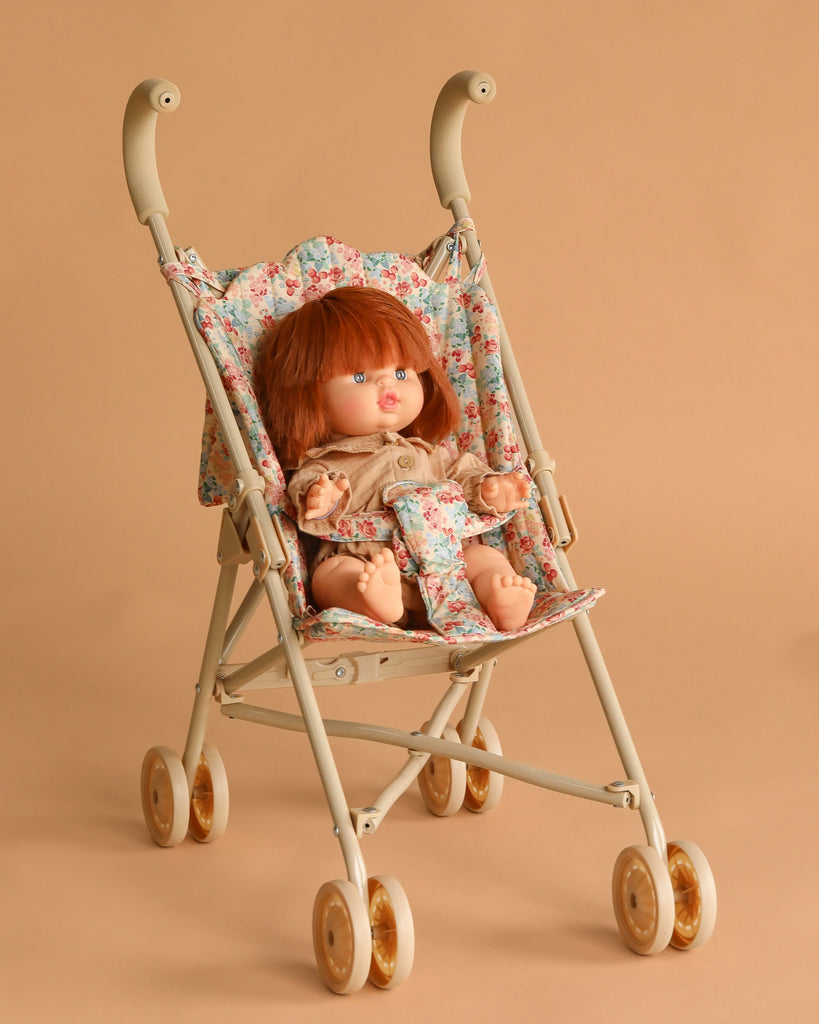 The Minikane Baby Doll (13") - Capucine with Clothing and Stroller Set features a red-haired doll in a beige outfit, seated in a floral-patterned stroller with beige wheels, set against a light brown background.