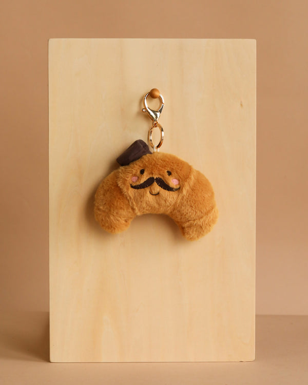 The Mon Ami - Monsieur Croissant Keychain / Bag Charm features a plush croissant with a cute face, rosy cheeks, brown mustache, and small hat—an adorable accessory for your keys or bag. Displayed on a light wooden board with a beige background.