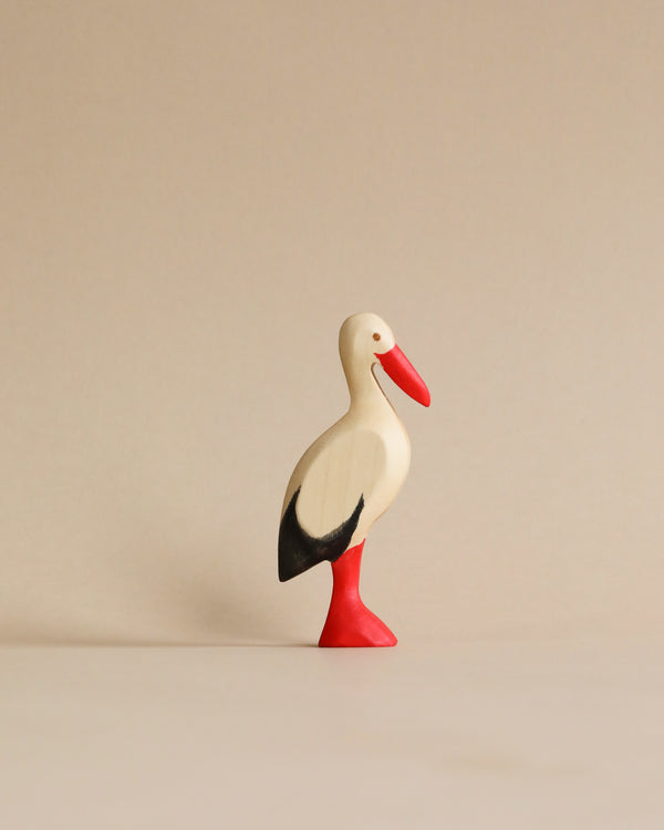 A Handmade Holzwald Stork figurine with a white body, black tail, and red beak and legs, standing upright on a beige background. This piece is an example of sustainable toys.