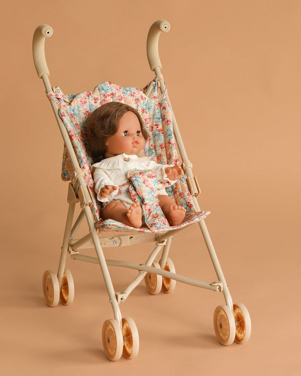 The Minikane Baby Doll (13") - Leopoldine with Clothing and Stroller Set features a brown-haired doll in a white outfit seated in a Konges Sloejd floral stroller with safety straps, against a light brown background.