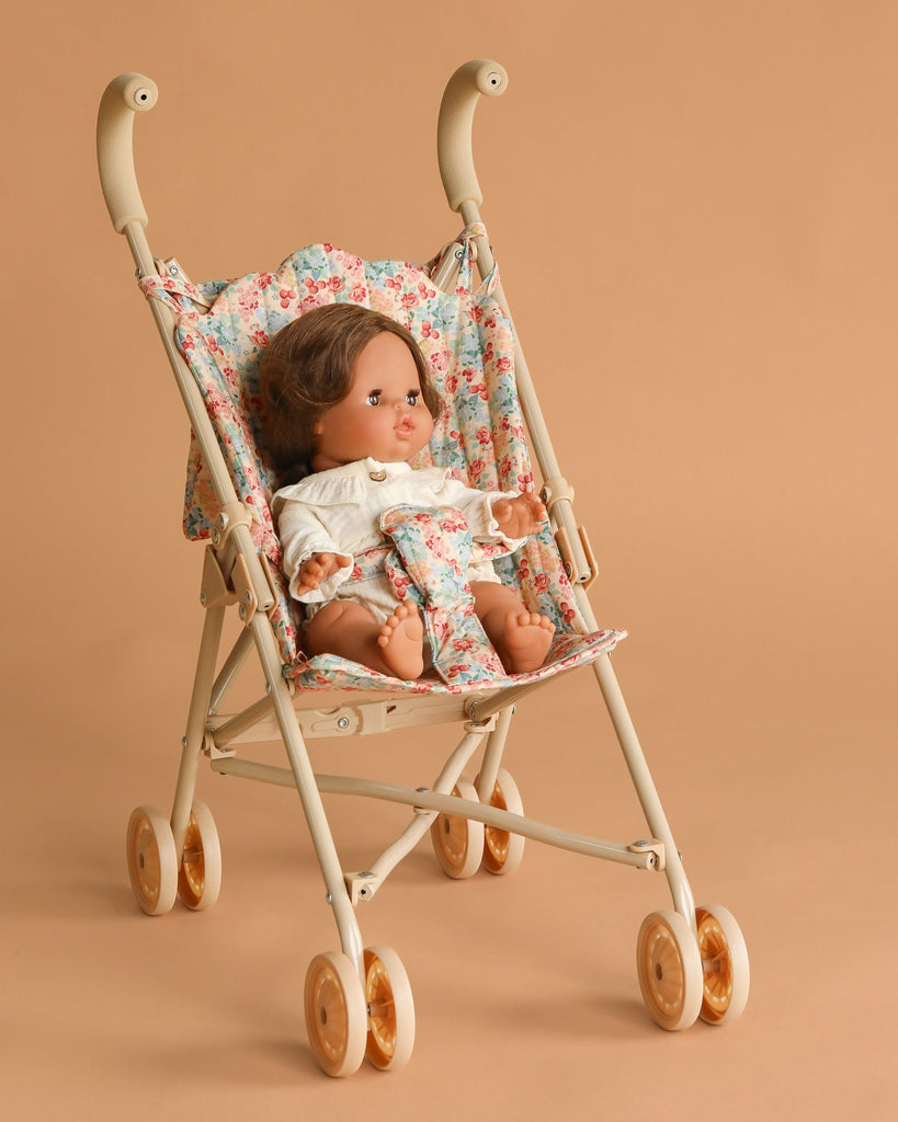 The Minikane Baby Doll (13") - Leopoldine with Clothing and Stroller Set features a brown-haired doll in a white outfit seated in a Konges Sloejd floral stroller with safety straps, against a light brown background.