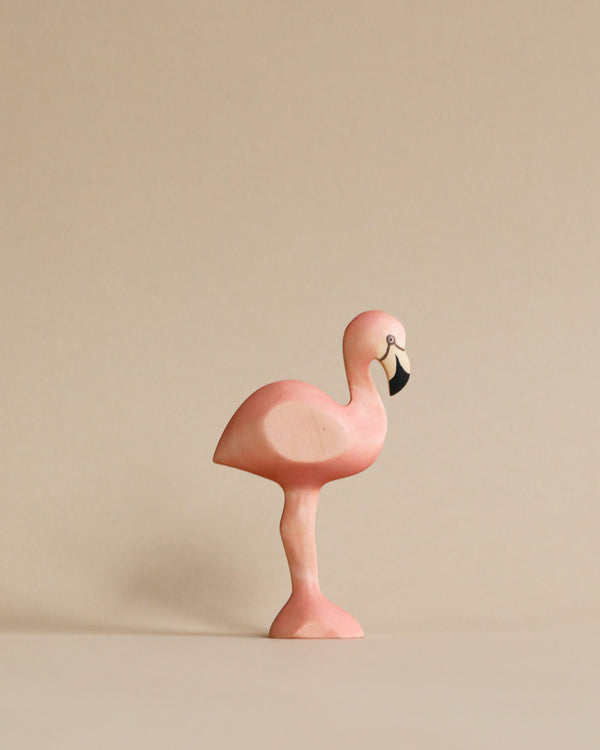 A Handmade Holzwald Flamingo from Holzwald wooden toys, with a smooth finish and a light pink color, stands against a neutral beige background, displaying its iconic one-legged pose.