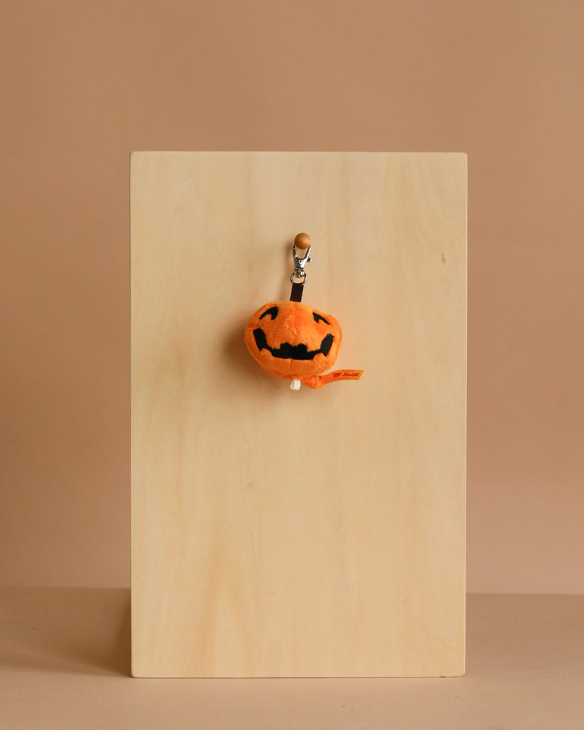The Steiff Little Pumpkin Keychain, a small plush pumpkin with a jack-o'-lantern face, hangs from a hook on a wooden board against a beige background—making it the perfect cute Halloween accessory.