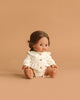 The Minikane Baby Doll (13") Leopoldine, with medium skin, brown hair and eyes, wears a cream buttoned outfit and sits against a plain tan background. This adorable anatomically correct doll is made in Spain.