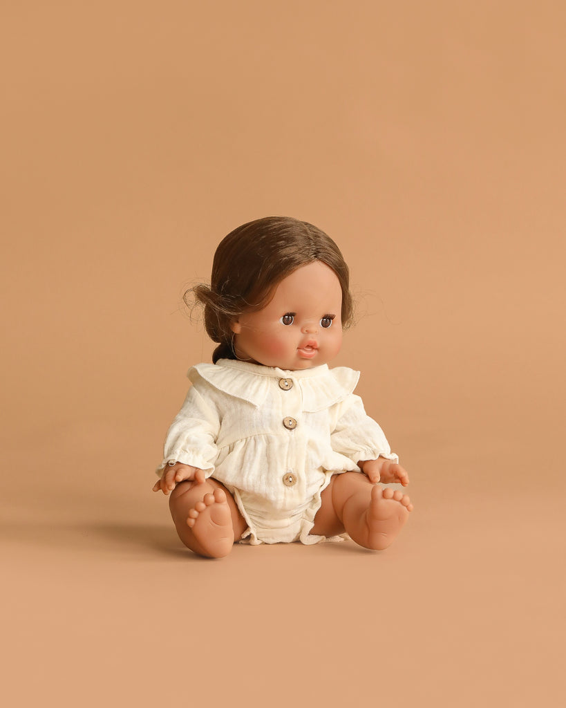 The Minikane Baby Doll (13") Leopoldine, with medium skin, brown hair and eyes, wears a cream buttoned outfit and sits against a plain tan background. This adorable anatomically correct doll is made in Spain.